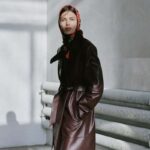 Magda Butrym x H&M. Zdjęcie przedstawia modelkę pozującą w projektach Magdy Butrym stworzonych we współpracy ze szwedzką sieciówką H&M. Kobieta ma na głowie zawiązaną chustkę w róże oraz skórzany brązowy płaszcz z wiązaniem w talii.