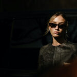 LVMH planuje podwyżki. Stylowa kobieta w czarnych okularach przeciwsłonecznych i eleganckiej, ciemnej marynarce z fakturą i zdobieniami, sfotografowana w półcieniu na tle ciemnego samochodu.