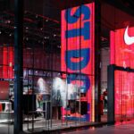 Zwolnienie w Nike. Zdjęcie przedstawia sklep amerykańskiej marki Nike, zostało zrobione nocą. Widać na nim ekrany z reklamą, charakterystyczne logo marki, a w tle – wieszaki z ubraniami sportowymi.