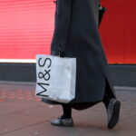 Kobieta idzie ulicą, trzymając w ręce białą papierową torbę z Marks & Spencer.