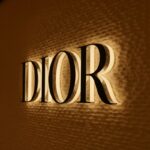 Szyld marki Dior – ciemny, podświetlony, znajdujący się strukturalnej ścianie. Po lewej stronie delikatnie widoczna szyba z kratami i manekinem.
