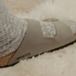Zbliżenie na szare zamszowe klapki Birkenstock, które wkrótce podrożeją. Buty są noszone z szarą skarpetką. Stopa stoi na białym futrzastym dywanie.