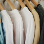 GAP wraca do Polski - otworzy pierwszy sklep stacjonarny w Westfield Arkadia. Na zdjęciu rząd kolorowych koszulek na drewnianych wieszakach wiszących w szafie, widocznych w zbliżeniu – w tym białe, niebieskie, beżowe i ciemnoszare ubrania.
