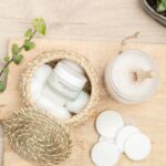 Naturalne produkty do pielęgnacji skóry na drewnianej desce: krem nawilżający w słoiczku w wiklinowym koszyku z płatkami kosmetycznymi i wacikami, słoik z solą do kąpieli i drewnianą łyżeczką, otoczony liśćmi bluszczu i doniczką z sukulentami.