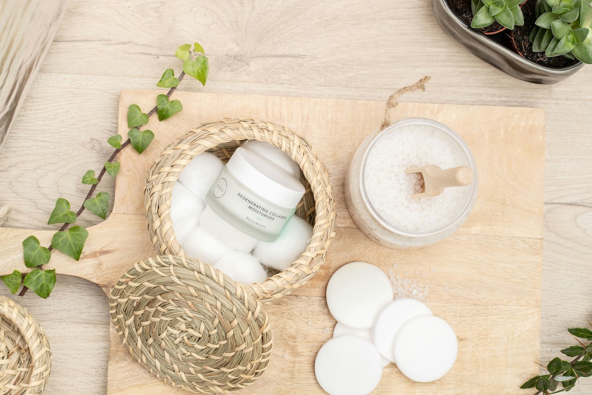 Naturalne produkty do pielęgnacji skóry na drewnianej desce: krem nawilżający w słoiczku w wiklinowym koszyku z płatkami kosmetycznymi i wacikami, słoik z solą do kąpieli i drewnianą łyżeczką, otoczony liśćmi bluszczu i doniczką z sukulentami.