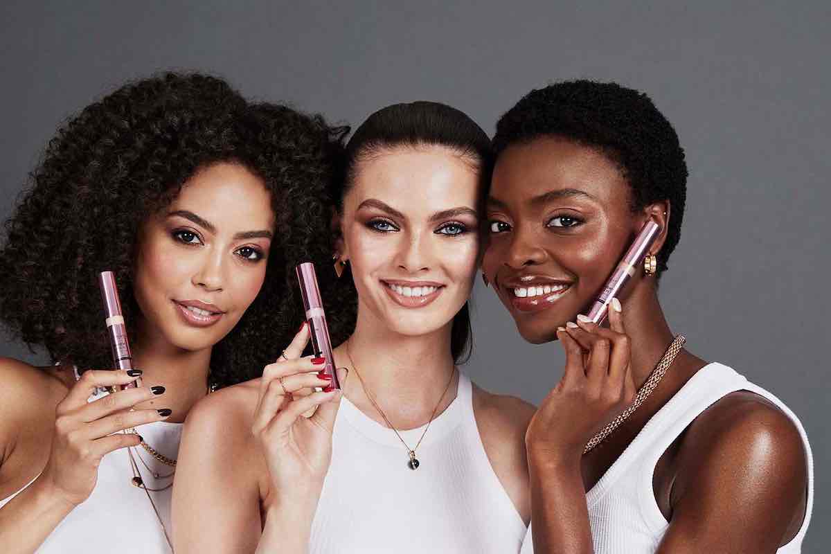 Marka Revolution Beauty na sprzedaż. Trzy uśmiechnięte kobiety o różnym kolorze skóry, ubrane w białe topy, prezentują tusze do rzęs Revolution Beauty w różowych opakowaniach.