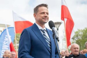 Kandydaci na prezydenta 2025 – styl i marki. Zdjęcie jednego z nich, Rafała Trzaskowskiego w granatowym garniturze, białej koszuli i krawacie w paski, stojącego na tle flag Polski.