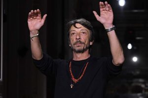 Pierpaolo Piccioli unoszący ręce po zakończonym pokazie. Włoski projektant ma na sobie czarną bluzkę, zegarek i czerwony wisiorek męski.