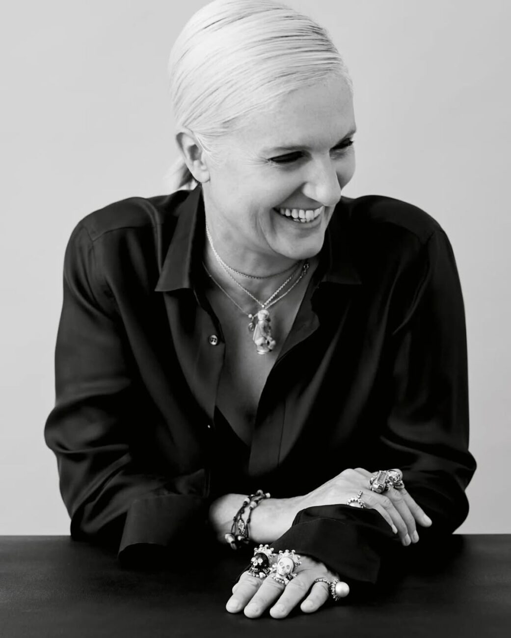 Maria Grazia Chiuri. Czarno-białe zdjęcie uśmiechniętej projektantki, pozującej w ciemnej koszuli z dużą liczbą biżuterii. Kobieta ma spięte blond włosy.