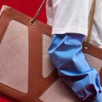 Zbliżenie na luksusową torbę Valentino w odcieniach brązu i beżu z łańcuszkiem na ramię, trzymaną przez modela w niebieskiej koszuli i białym T-shircie na czerwonym tle.