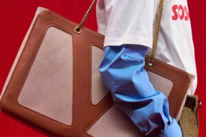 Zbliżenie na luksusową torbę Valentino w odcieniach brązu i beżu z łańcuszkiem na ramię, trzymaną przez modela w niebieskiej koszuli i białym T-shircie na czerwonym tle.