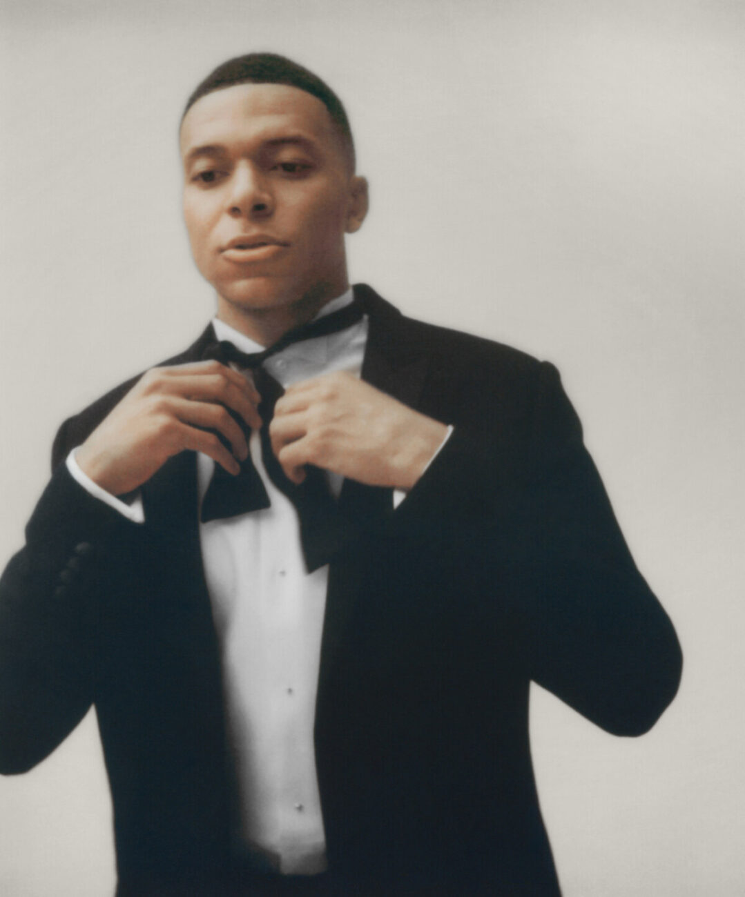 Kylian Mbappé z białej koszuli i czarnej marynarce, wiąże czarną muszkę. Zdjęcie zrobione w ruchu, to kadr filmu promocyjnego.