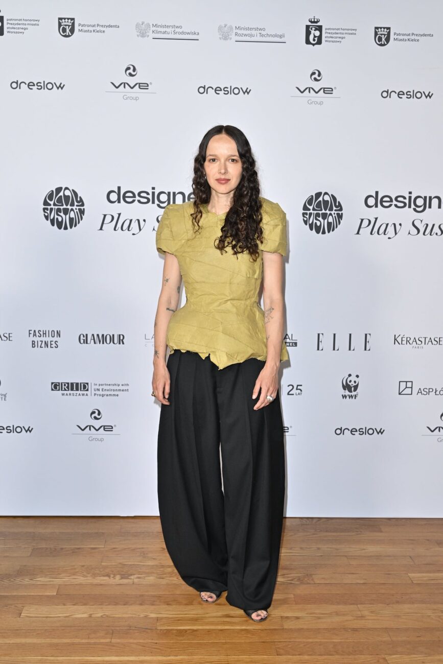 Monika Brodka w zielonej, asymetrycznej bluzce i szerokich czarnych spodniach pozuje na tle ścianki z logotypami partnerów wydarzenia "Designers Play Sustain".