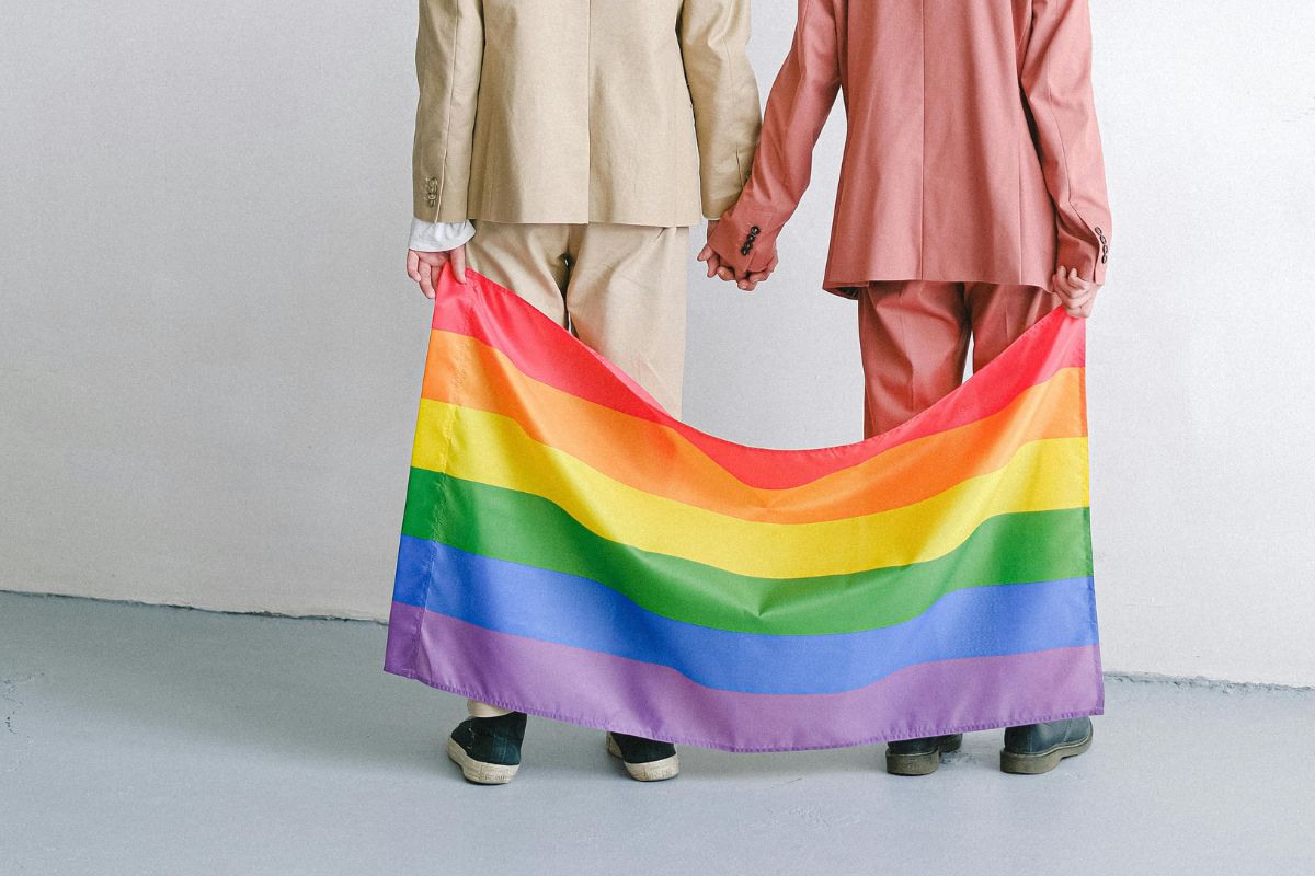 Zdjęcie przedstawiające marki na Pride Month. Widać dwóch mężczyzn w garniturach – jeden w beżowym, drugi w różowym – stoją tyłem, trzymają się za ręce i trzymają w ręce flagę tęczową.
