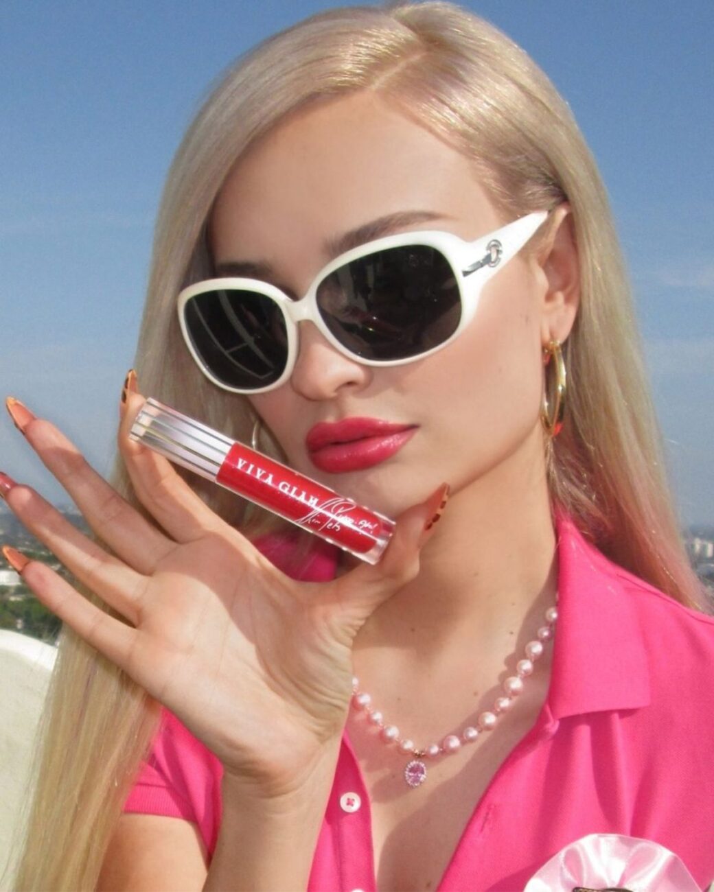 Zdjęcie przedstawiające kampanię Mac Viva Glam” Pride 2025 Collection z artystką Kim Petras. Razem stworzyli błyszczyk do ust „Viva Glam KIMmitment Lipglass Air” specjalnie na Pride Month 2025. Na zdjęciu znajduje się Petras, w długich blond włosach, różowej bluzce z kołnierzykiem, sznurem pereł na szyi, białych okularach. W ręce trzyma błyszczyk.