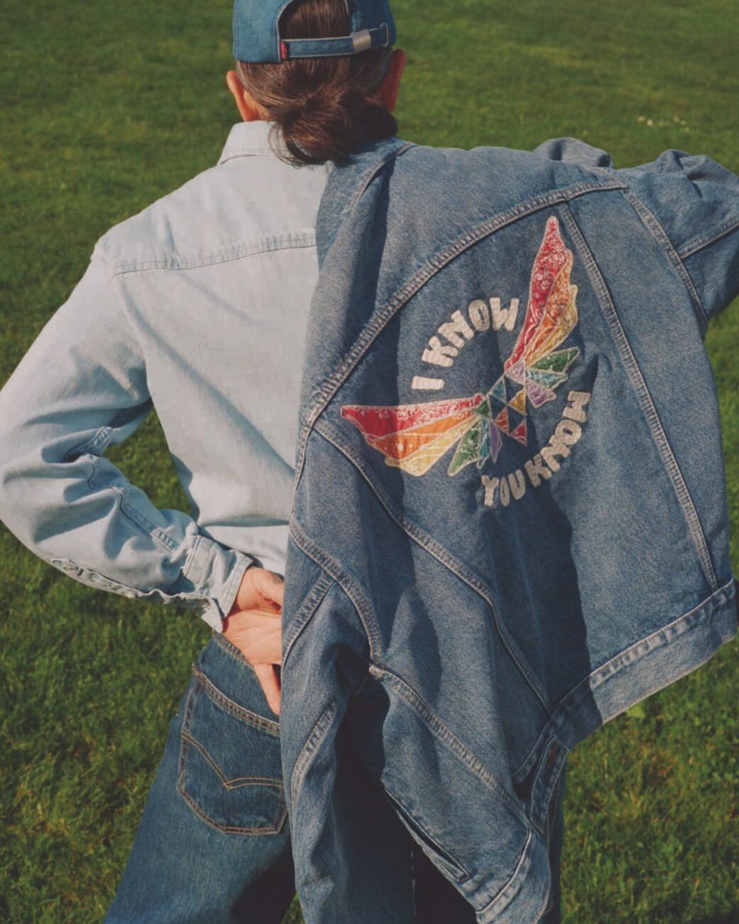 Zdjęcie przedstawiające kammapnię marki Levi's® Pride 2025 „Meet You In The Park”. Na zdjęciu stoi tyłem mężczyzn ubrany w jeansową koszulę, spodnie, czapkę z daszkiem oraz katanę z nadrukiem z tęczowymi skrzydłami.