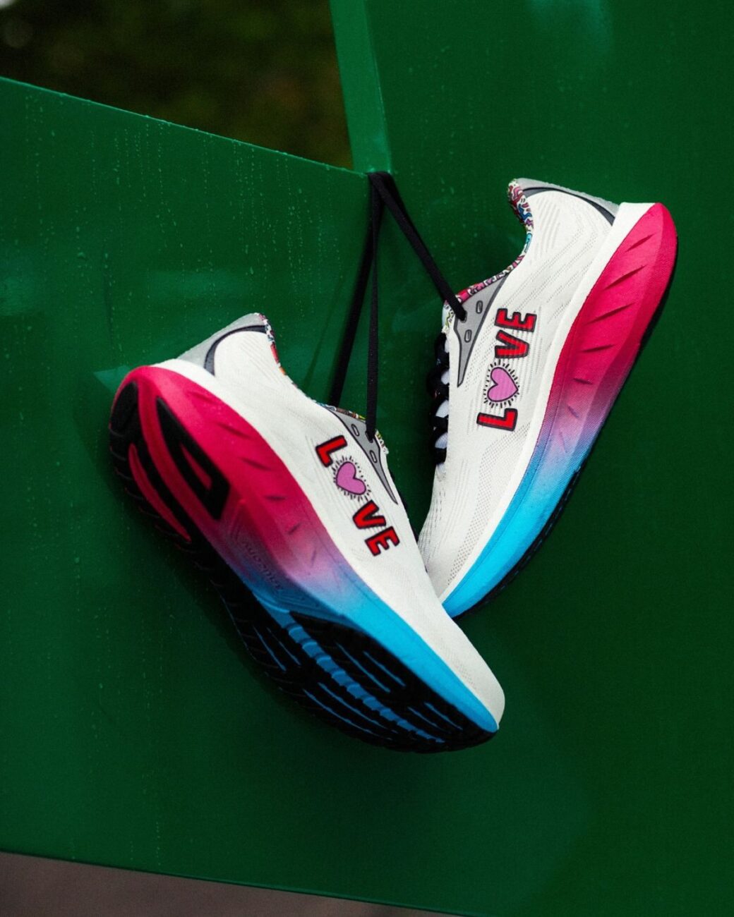 Zdjęcie przedstawiające Saucony i kolekcję Pride Month. Widać na nim sneakersy w kolorze białym z różową podeszwą i napisem Love. Są powieszone za sznurówki.