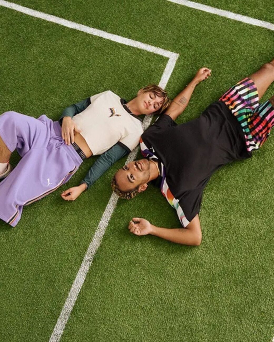Zdjęcie przedstawiające kampanię Puma „Equal Love Confederation”, czyli marki na Pride Month 2025, która przygotowała specjalną kolekcję. Na zdjęciu widać modeli, dziewczynę i chłopaka, ubranych w koszulki i spodnie z kolekcji. Leżą na murawie boiska.