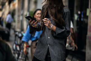 Asos blokuje konta w związku z nadmierymi zwrotami. Kobieta na ulicy w Mediolanie trzymająca smartfon i szminkę, ubrana w ciemnoszarą koszulę z długim rękawem i ozdobiona licznymi srebrnymi pierścionkami oraz bransoletkami; jej długie włosy częściowo zasłaniają twarz, a w tle widać innych przechodniów i zaparkowane rowery.