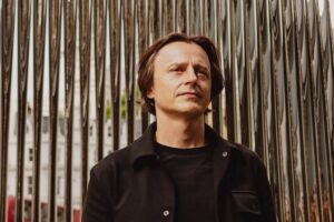 Oskar Zięta kim jest? Zdjęcie projektanta ubranego w czarny T-shirt i kurtkę, stoi na tle swojej rzeźby wykonanej w technologii FIDU.