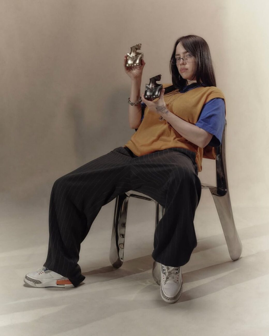 Zdjęcie Billie Eilish promującej swoje perfumy: jest ubrana w szerokie, prążkowane spodnie, pomarańczowy T-shirt i sneakersy. Siedzi na krześle Chippensteel Oskara Zięty i trzyma w rękach perfumy.