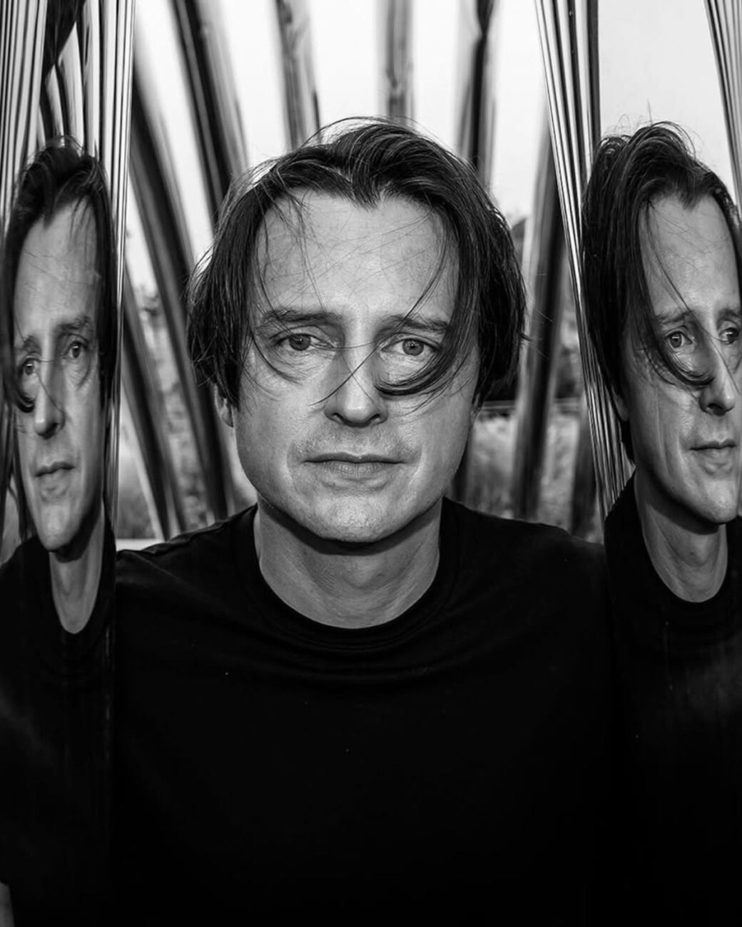 Zdjęcie Oskara Zięy w czarno-białej wersji. To portret. Stoi przy swojej rzeźbie NAWA we Wrocławiu na Wyspie Słodowej. Stoi na wprost obiektywu i odbija się na ścianach swojej lśniącej rzeźby.