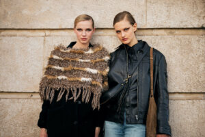 Francja wprowadza zakaz fast fashion. Na zdjęciu dwie kobiety stojące obok siebie na tle kamiennej ściany. Po lewej kobieta w czarnym płaszczu i narzutce z brązowego futra w paski, z zaczesanymi do tyłu blond włosami i mocnym makijażem oczu. Po prawej kobieta w czarnej skórzanej kurtce, dżinsach i torbie na ramię, również z zaczesanymi włosami i poważnym wyrazem twarzy.