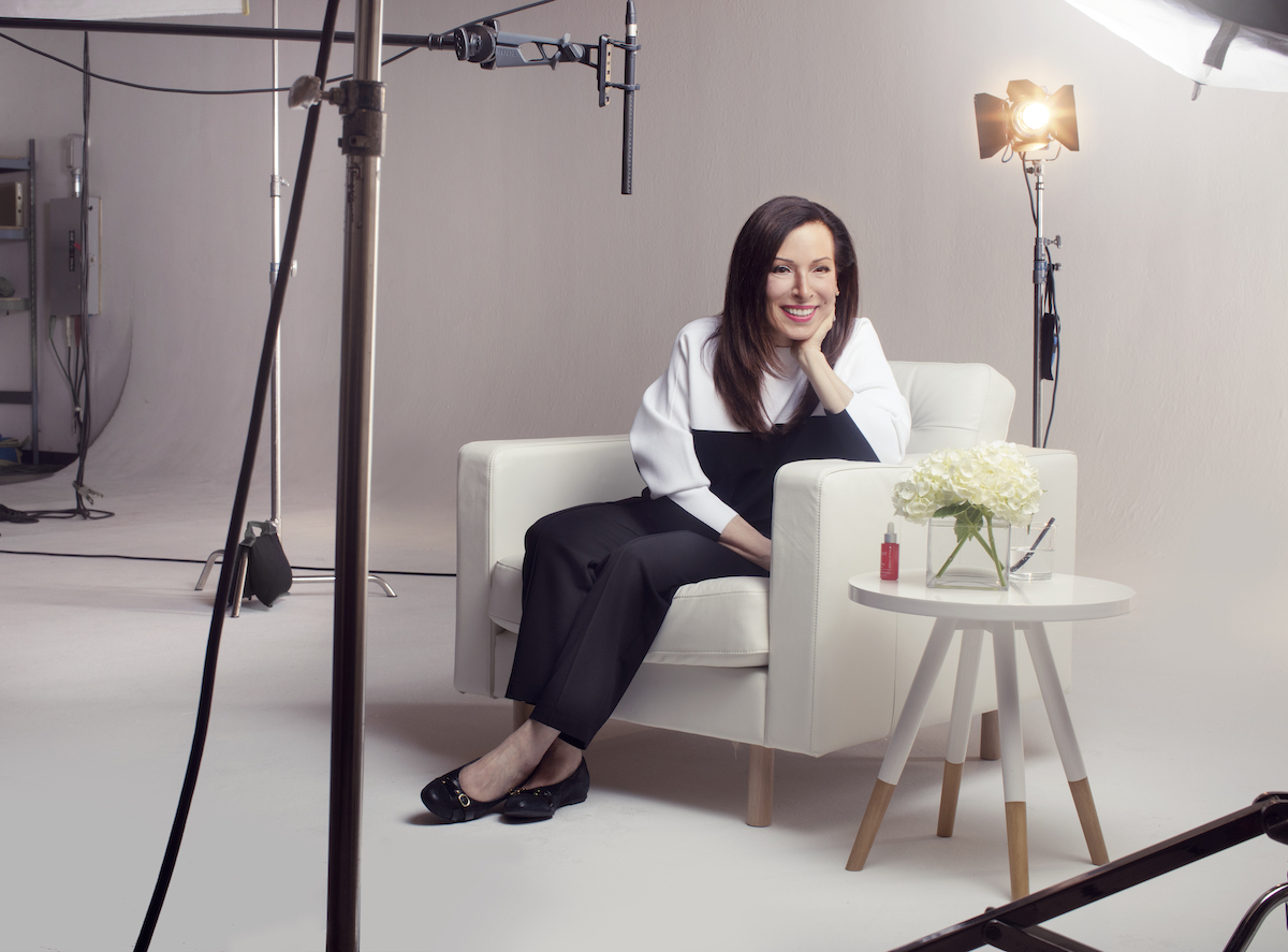Kobieta siedząca na białym fotelu w profesjonalnym studiu fotograficznym, uśmiechnięta, z długimi ciemnymi włosami, ubrana w biało-czarny strój. Obok niej biały stolik z kwiatami i kosmetykiem.