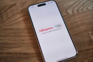 Zdjęcie telefonu leżącego na drewnianym blacie. Na białym ekranie wyświetla się nazwa platformy AliExpress, która według Komisji Europejska narusza prawo. Oprócz tego na ekranie widać logo Igrzysk Olimpijskich oraz napis "Official E-commerce Services Partner".