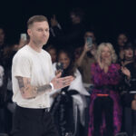 Projektant Matthew M. Williams po zakończonym pokazie dla Givenchy. Mężczyzna ma wytatuowane ramiona, nosi biały T-shirt i czarne spodnie z paskiem. W tle widać widownię pokazu bijącą brawo.
