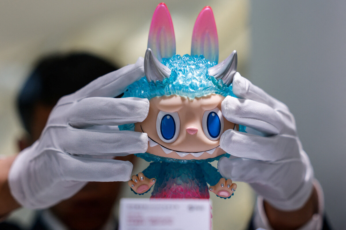 Pracownik prezentuje figurkę Labubu na wystawie przed Yongle International Auction w Pekinie. Pracownik ma białe rękawiczki, podczas gdy figurka jest w intensywnie niebieskim kolorze z różowymi akcentami. Ma też niebieskie oczy i rogi.
