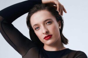 Iga Świątek na pierwszym miejscu rankingu Forbes Women - 100 Najcenniejszych Kobiecych Marek Osobistych 2025. Na zdjęciu Iga Świątek w kampanii Lancome, ubrana w czarne dopasowane body. Rękę trzyma na głowie. Ma czerwone usta i delikatny makijaż.
