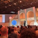Scena wydarzenia LVMH Innovation Award Ceremony, z elegancką dekoracją w ciepłych odcieniach pomarańczu i beżu. Na scenie stoi kobieta w ciemnym garniturze, prowadząca wydarzenie, a jej obraz jest również wyświetlany na dużym ekranie z boku sceny. Przed sceną siedzi liczna publiczność, a na tle znajduje się napis "LVMH Innovation Award Ceremony". W tle widoczne są także logotypy "Viva Technology".