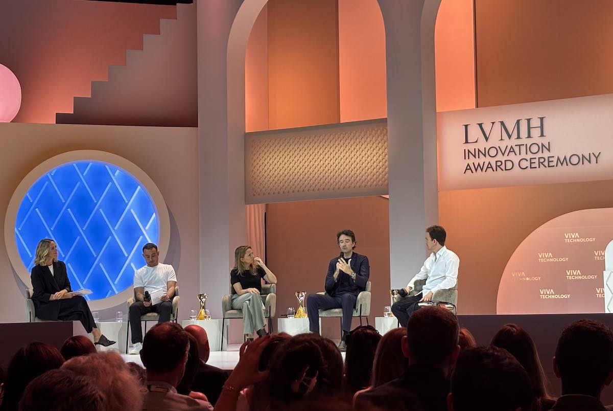 Panel dyskusyjny podczas ceremonii wręczenia nagród LVMH Innovation Award. Na scenie siedzi pięć osób, w tym trzy mężczyzn i dwie kobiety, rozmawiających w eleganckim otoczeniu z ciepłym oświetleniem w odcieniach pomarańczowego i beżowego. Za nimi widnieje napis „LVMH Innovation Award Ceremony” oraz logotypy „Viva Technology”. Na stolikach obok panelistów stoją złote trofea. W tle znajduje się dekoracja z łukami i dużym, niebieskim, podświetlonym okręgiem.