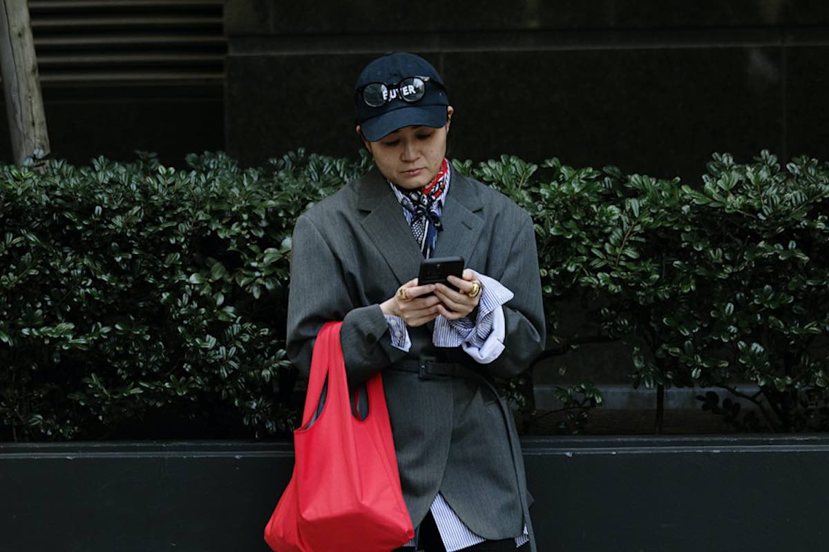 Osoba w czapce z daszkiem i szarym, oversizowym płaszczu, z czerwonym materiałowym workiem, patrzy w telefon.