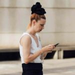 Whatsapp reklamy. Wielka zmiana na najpopularniejszym komunikatorze świata. Na zdjęciu kobieta z telefonem w rękach, z dużą czarną gumką na środku głowy i w białym topie bez rękawów.