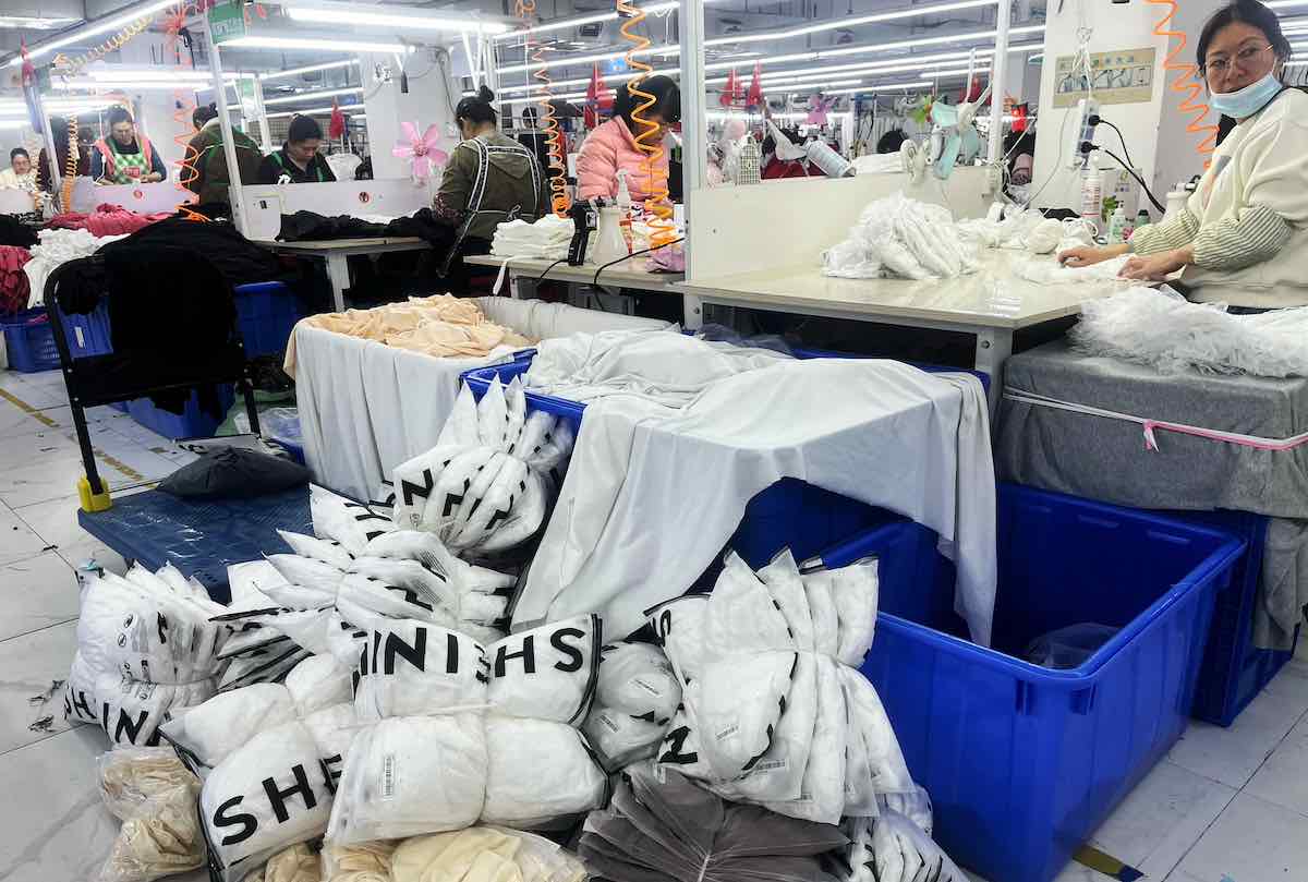 Shein produkcja Indie. Zdjęcie przedstawia halę produkcyjną w fabryce odzieżowej, gdzie pracownicy szyją i pakują ubrania. Na pierwszym planie widać stosy plastikowych toreb oznaczonych logotypem „SHEIN” wypełnionych białymi i kremowymi ubraniami. W tle widoczne są stanowiska pracy z maszynami do szycia, przy których siedzą pracownicy w odzieży roboczej.