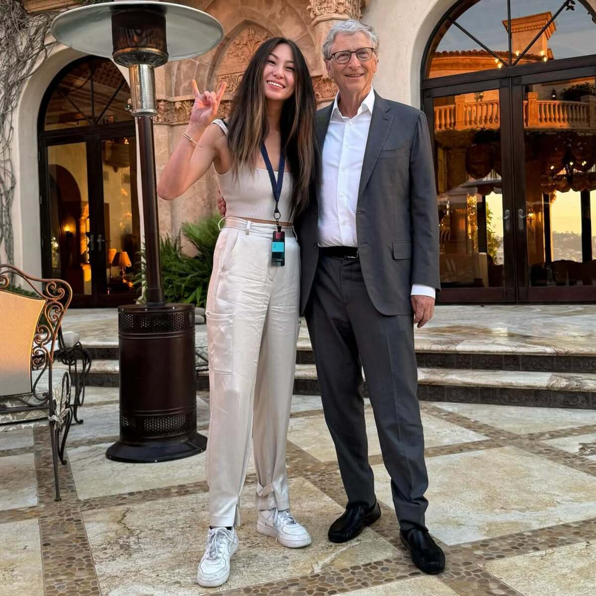 Zdjęcie najmłodszej self-made miliarderki na świecie z Billem Gatesem. Lucy Guo (po lewej) pozuje w rozpuszczonych włosach, w jasnych spodniach i białych butach sportowych. Wykonuje znak pokoju. Z kolei Bill Gates (po prawej) uśmiecha się i ma na sobie ciemnoszary garnitur, białą koszulę, czarne eleganckie buty i okulary.
