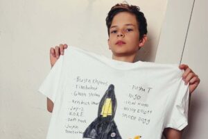 Zdjęcie 11-letniego Dylana, założyciela marki Dylan’s T-shirt Club. Chłopiec prezentuje własnoręcznie zrobioną białą koszulkę napisami i rysunkiem.