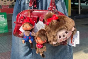Czy Monchhichi dorówna popularnością Labubu? Zdjęcie czerwonej torebki trzymanej przez kobietę w jeansach. Na torebce są zawieszone 3 pluszowe zabawki z metkami. To Monchhichi: małpki w niebieskich ogrodniczkach, czerwonym śliniaczku oraz duża głowa z kokardką.