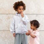 H&M Adorables. Zdjęcie reklamowe dwóch dzieci – starszego, wyższego chłopca w jasnej kurtce sportowej i luźnych spodniach oraz dziewczynki w różowej sukience. Dzieci mają ciemne kręcone włosy.