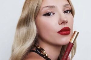 Coty – sprzedaż marek. Zdjęcie modelki w rozpuszczonych blond włosach, reklamującej czerwony błyszczyk lub pomadkę. Kobieta ma pomalowane nią usta i makijaż oczu w stylu kociego oka.