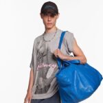 Zdjęcie modela pozującego w ubraniach z kolekcji Balenciaga x Britney Spears. Mężczyzna nosi szary T-shirt ze zdjęciem piosenkarki i czapkę z limitowanej kolekcji. Na ramieniu ma dużą niebieską, wypakowaną torbę.