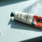 Zdjęcie kremu do rąk The Body Shop. Kosmetyk leży na oświetlonym słońcem blacie, ma zieloną zakrętkę, a na tubce widać napis "Strawberry". Obok kremu leży złota biżuteria – kolczyki i łańcuszek albo bransoletka.