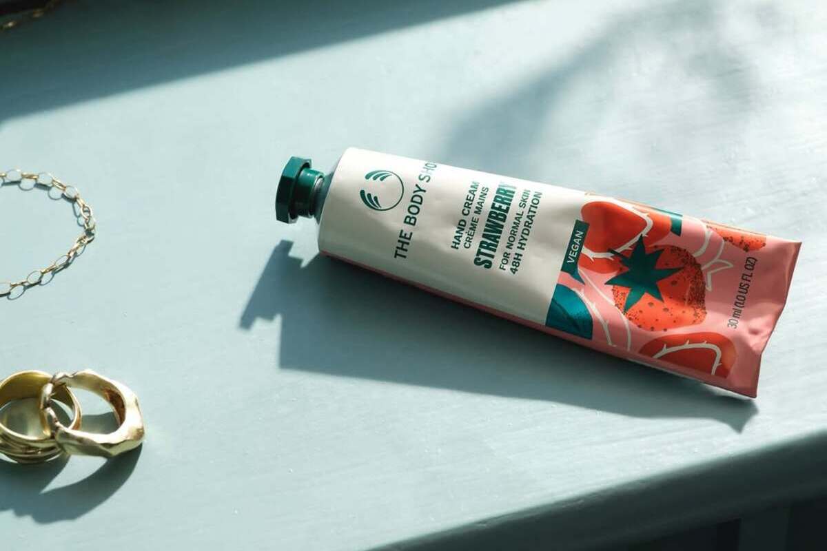 Zdjęcie kremu do rąk The Body Shop. Kosmetyk leży na oświetlonym słońcem blacie, ma zieloną zakrętkę, a na tubce widać napis "Strawberry". Obok kremu leży złota biżuteria – kolczyki i łańcuszek albo bransoletka.
