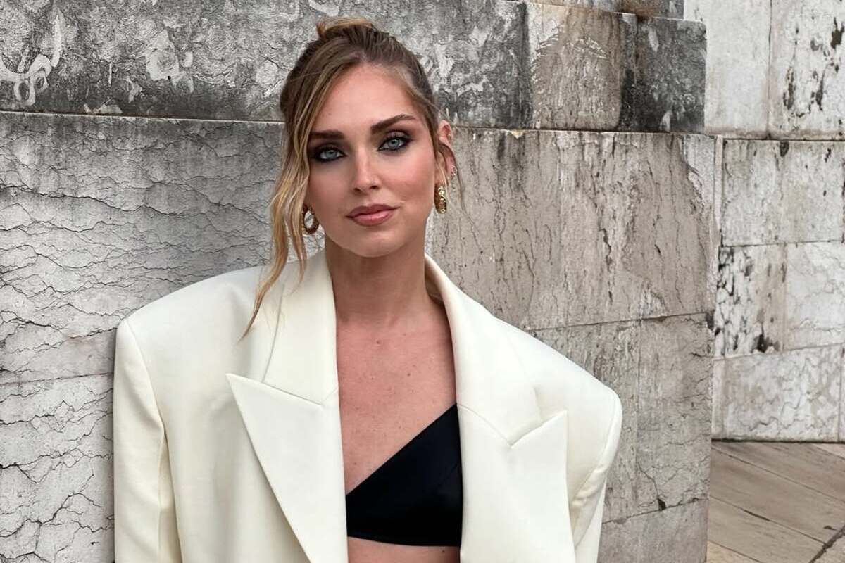 Chiara Ferragni likwiduje firmę. Zdjęcie włoskiej influencerki w spiętych włosach z luźno puszczonym pasemkiem przy twarzy i mocnym makijażu wieczorowym. Kobieta nosi kremową marynarkę o mocno oversizowym kroju. Pod nią ma czarny stanik. Stoi na tle muru.