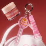 Zdjęcie reklamowe kolekcji H&M Beauty i Chupa Chups, która trafi niedługo do sklepów. NA zdjęciu kosmetyk w małym słoiczku z jasną zakrętką, breloczek w kształcie lizaka z różowym błyszczykiem oraz część torebki.