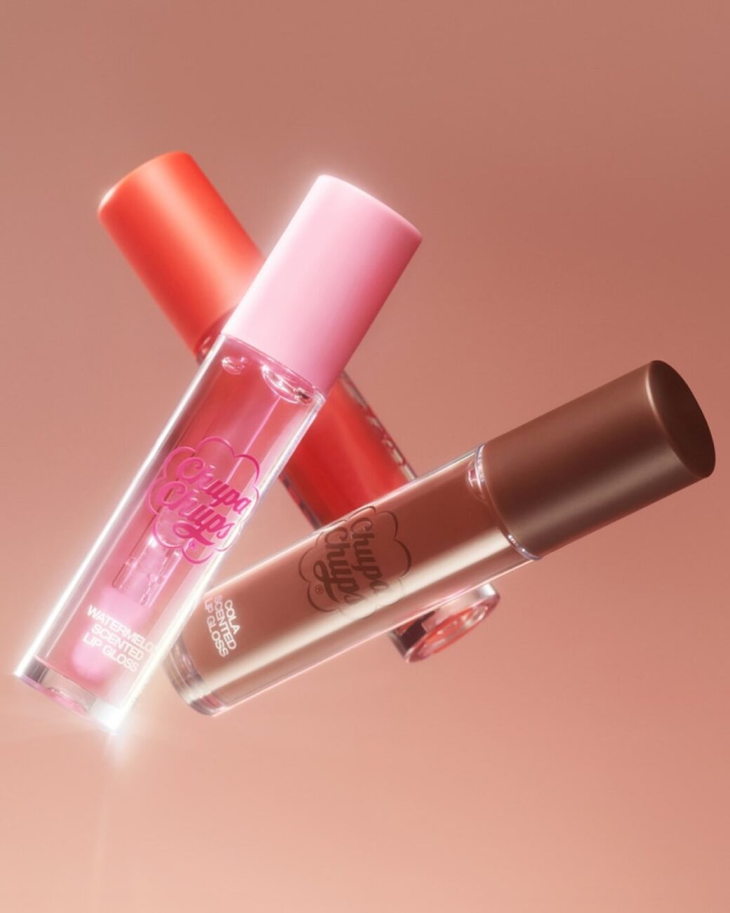 Zdjęcie 3 błyszczyków z kolekcji H&M Beauty i Chupa Chups. Kosmetyki mają różne kolory: różowy, czerwony i brązowy. Unoszą się w powietrzu.