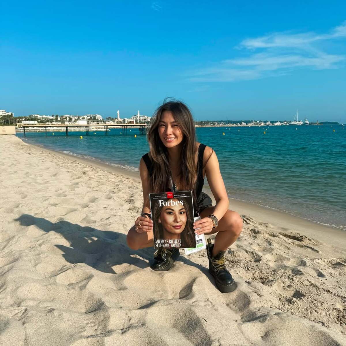 Najmłodsza miliarderka na świecie, Lucy Guo, pozuje na plaży z magazynem Forbes. Kobieta jest na okładce czasopisma. Lucy Guo uśmiecha się, ma rozpuszczone włosy oraz nosi masywne czarne buty.