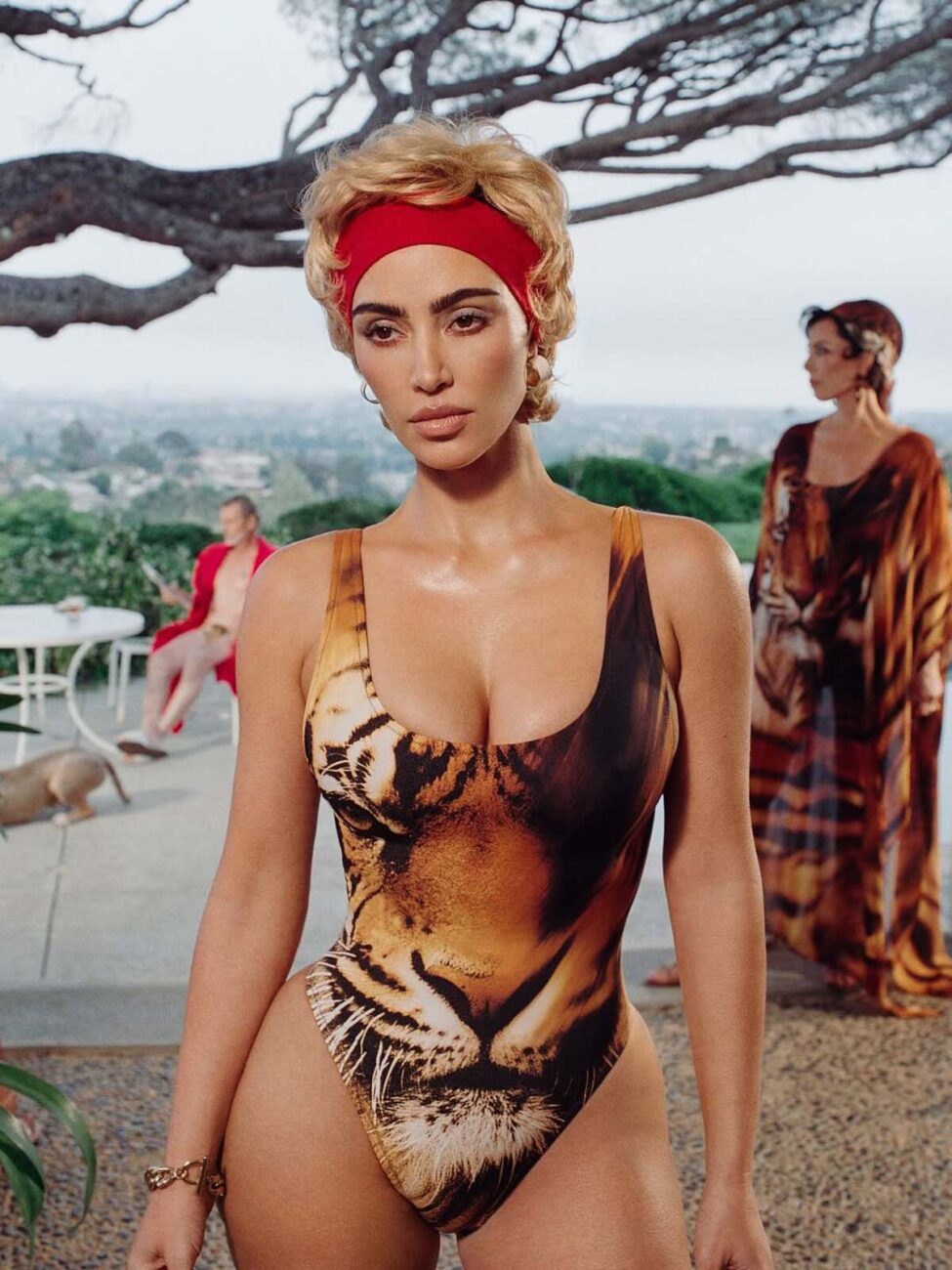 Kim Kardashian w krótkiej blond peruce, pozuje w stroju kąpielowym z nadrukiem tygrysa i czerwonej opasce. To zdjęcie z kampanii reklamowej kolekcji Roberto Cavalli x SKIMS.